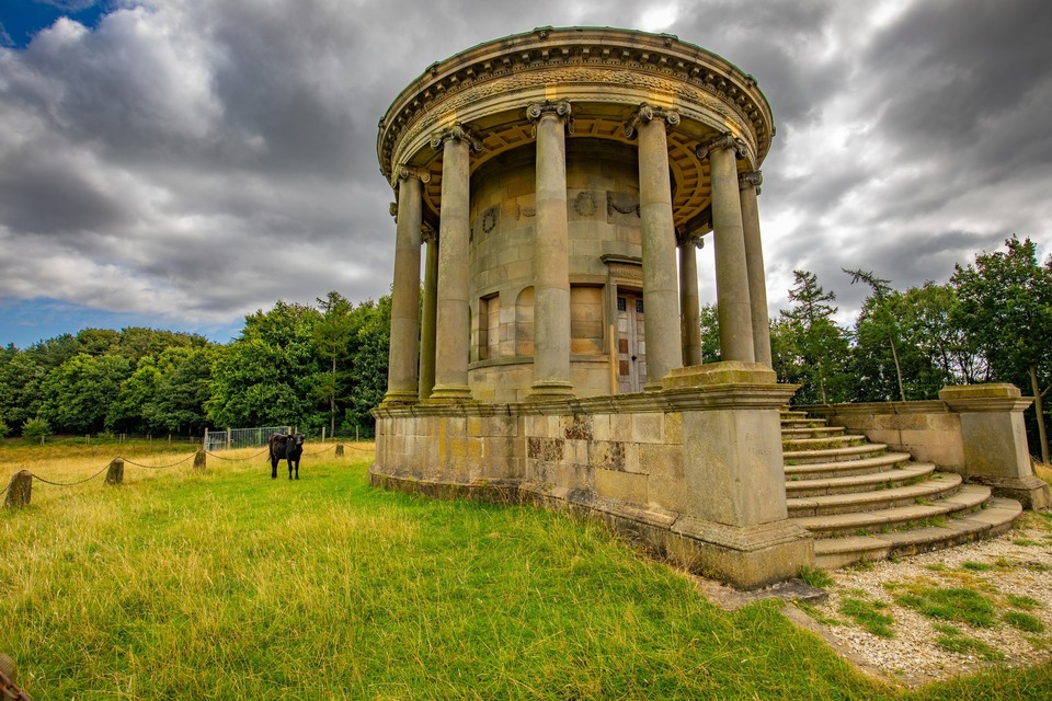 Circular stone structure with classical columns, resembling a temple or rotunda, set in a grassy field with a black cow nearby and trees in the background under a cloudy sky.